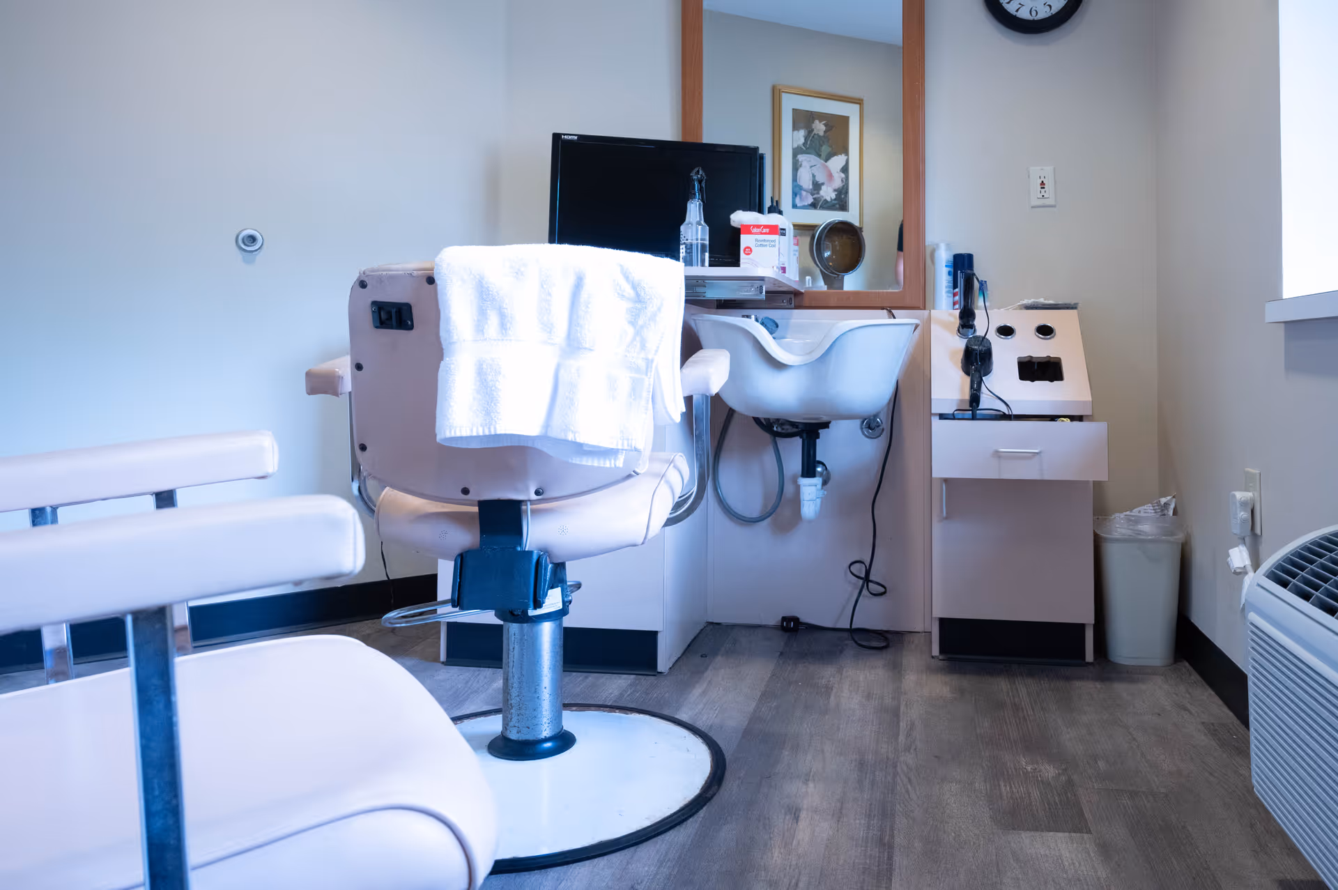 Interior view of a salon or grooming area with a beige salon chair draped with a white towel, a small sink, a mirror, and various grooming supplies on the counter. The room has light-colored walls and wood flooring.
