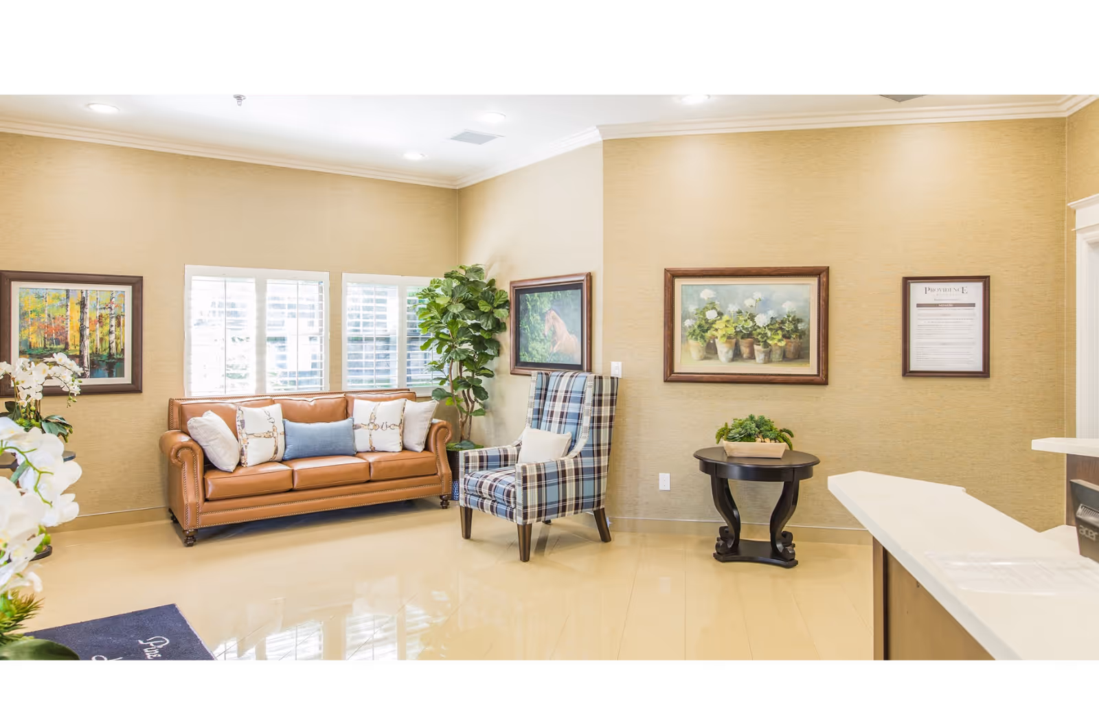 Bright reception seating area with a leather sofa, plaid armchair, framed artwork, and a small table in a senior living facility lobby.