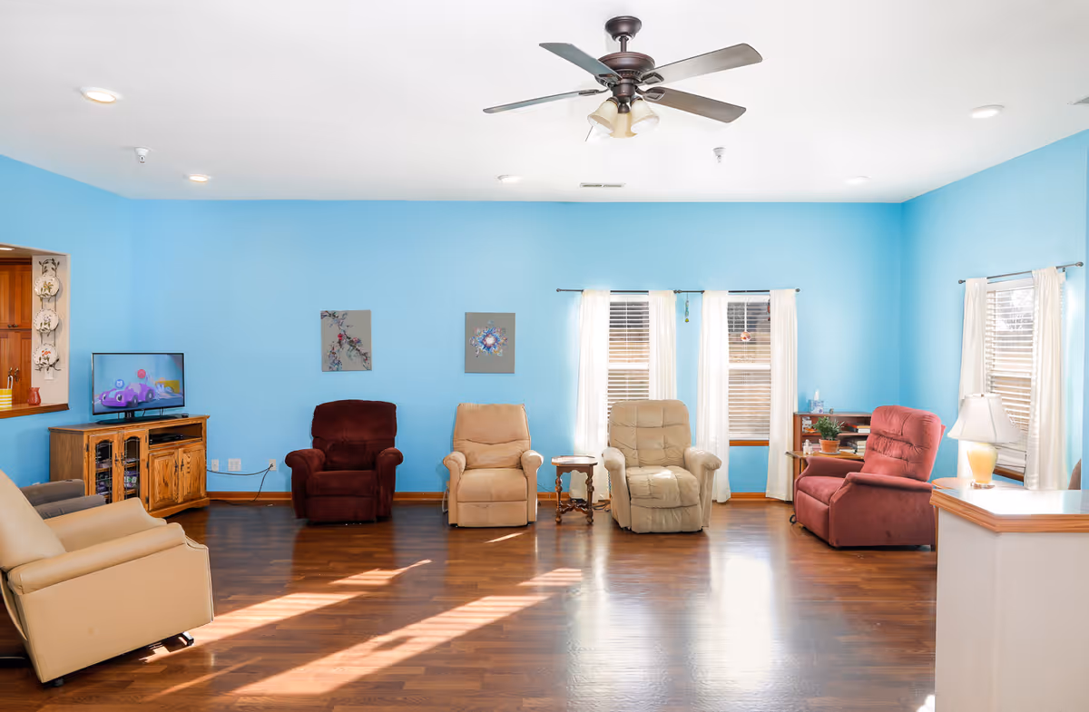 Bright communal living room with several recliner chairs arranged along a light blue wall, a TV cabinet, windows with curtains, and a ceiling fan.