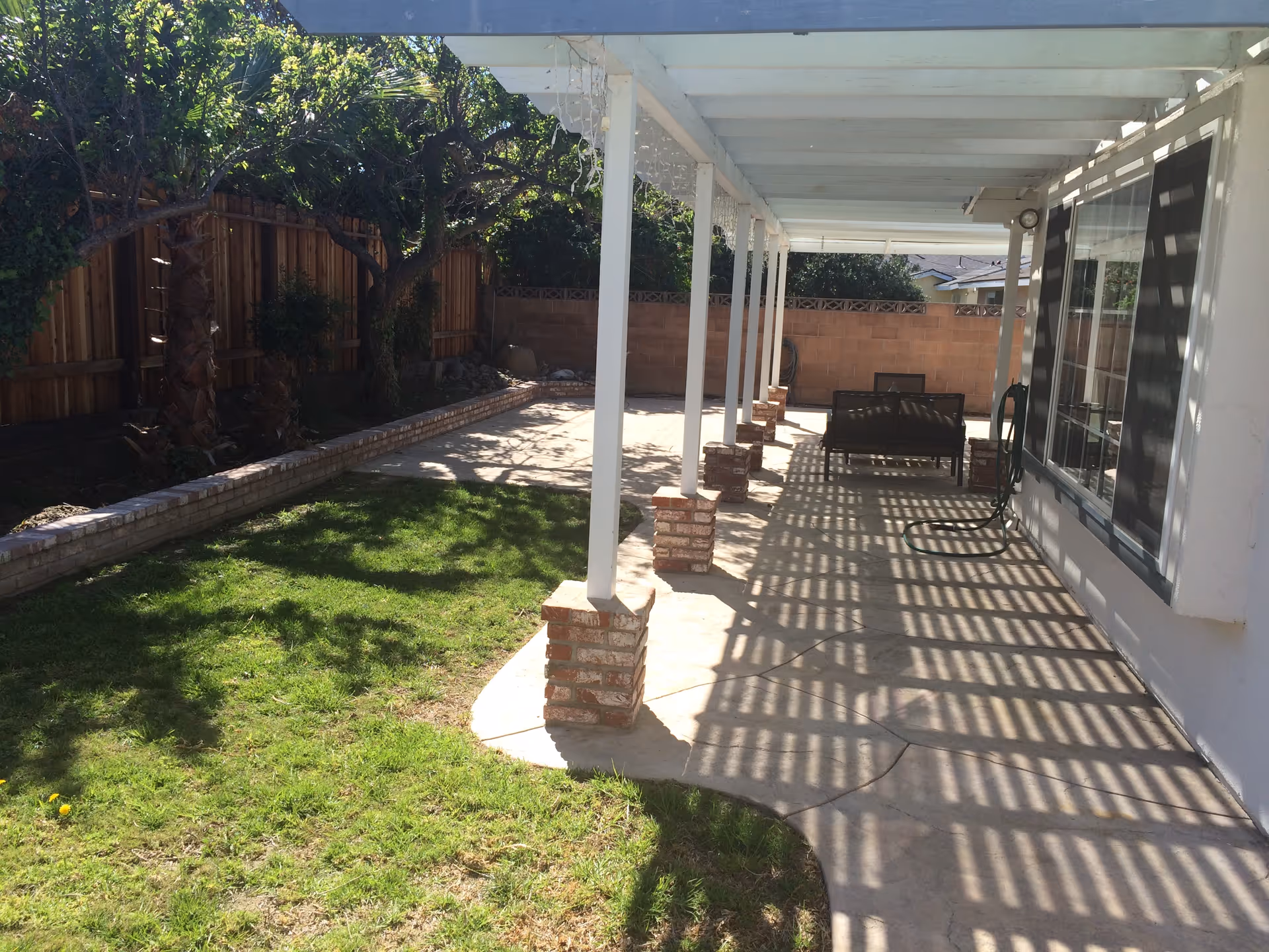 A backyard patio area with a covered walkway supported by white posts with brick bases. The patio has concrete flooring and outdoor seating. There is a grassy lawn area to the left, bordered by a wooden fence and trees providing shade.