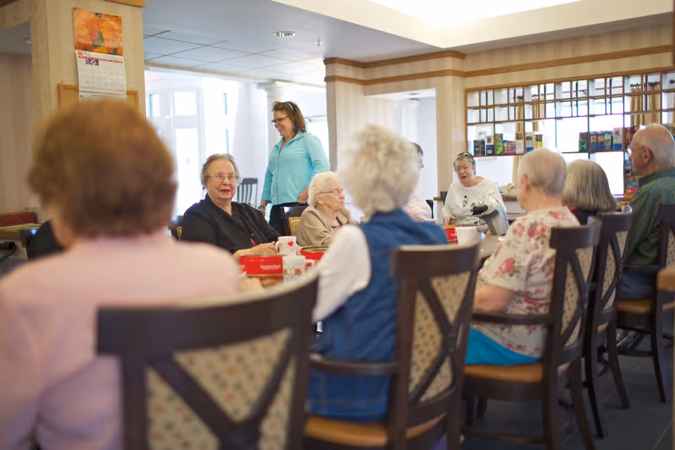 A group of elderly people sitting around a large table in a well-lit room, engaging in conversation. A caregiver or staff member stands nearby smiling. The room has wooden chairs with patterned cushions and a calendar on the wall.