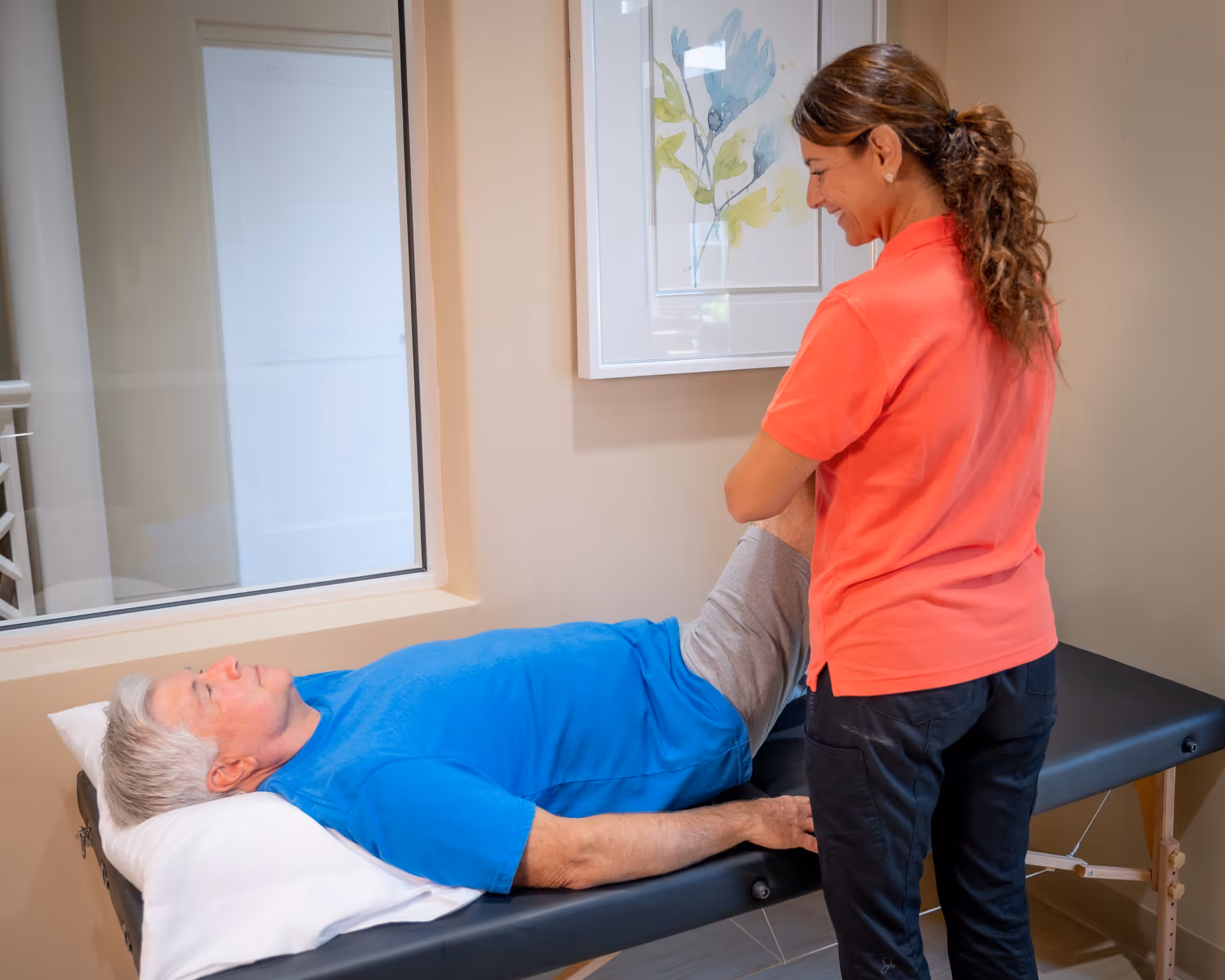 A healthcare professional in a coral shirt assists an elderly man lying on a treatment table by gently holding and stretching his leg in a well-lit room with a window and framed artwork on the wall.