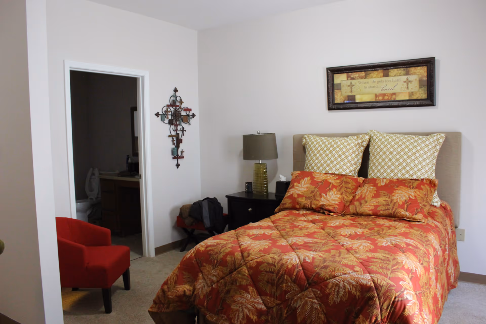 A bedroom with a bed covered in a red and orange leaf-patterned comforter and matching pillows. There is a nightstand with a lamp and tissues next to the bed. A framed inspirational quote hangs above the bed. To the left, there is a red armchair and an open doorway leading to a bathroom with a visible toilet and sink. A decorative wall cross is mounted on the wall near the bathroom door.