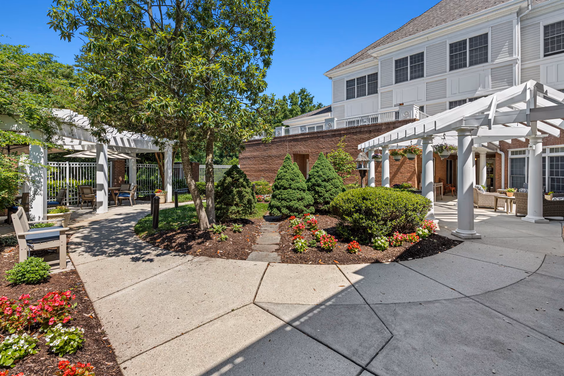 Sunny landscaped courtyard with pergolas, seating, walkways, and flower beds in front of a multi-story building.