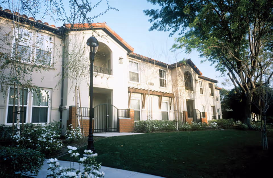 Exterior view of a two-story residential building with white walls and a red-tiled roof, surrounded by trees and greenery. There is a black street lamp and a gated entrance visible in front of the building.