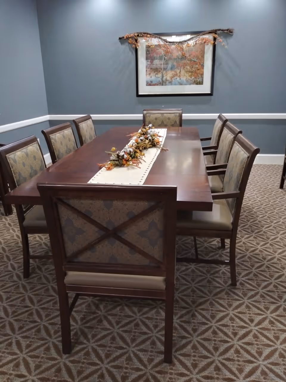 A dining room with a rectangular wooden table surrounded by eight upholstered chairs. The table has a decorative runner with a floral centerpiece. The walls are painted blue-gray and feature a framed artwork with autumn leaves and a garland hanging above it. The floor is carpeted with a patterned design.