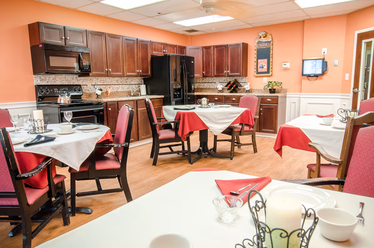 A dining room area with several tables covered in white and red tablecloths, set with plates, bowls, glasses, and utensils. The room features wooden chairs with red cushions, a kitchen area with dark wooden cabinets, a black refrigerator, stove, microwave, and a tiled backsplash. The walls are painted peach with white wainscoting, and there is a ceiling fan and overhead lighting.