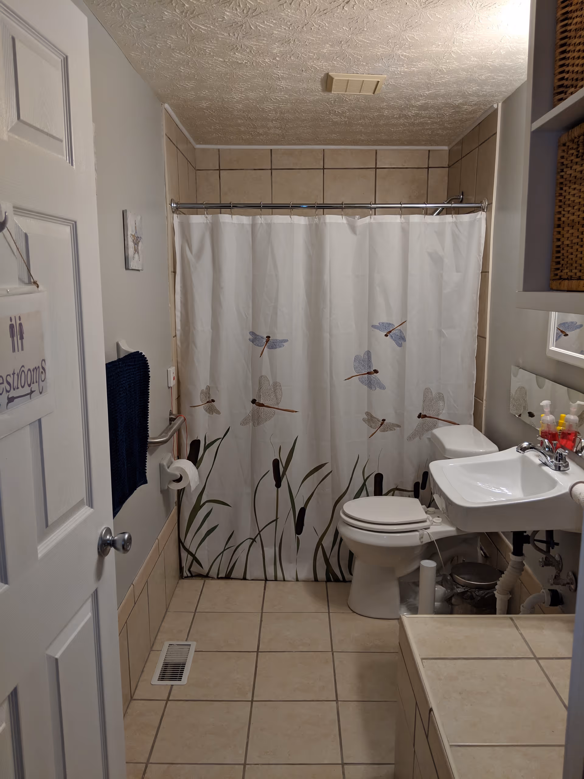 A clean bathroom with beige tiled floor and walls. There is a white shower curtain decorated with dragonflies and cattails, a white toilet, and a white sink with a mirror above it. A blue towel hangs on the wall next to the toilet paper holder. The door has a sign that reads 'Restrooms'.