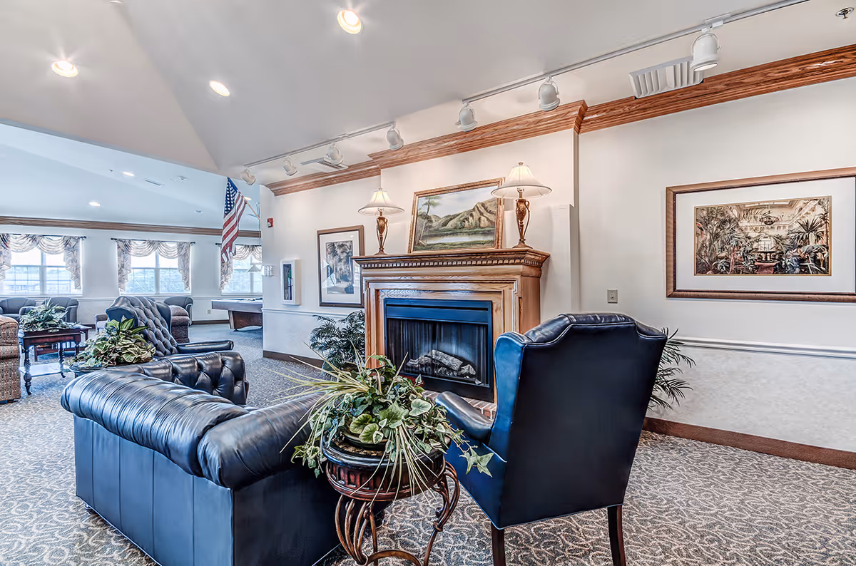A bright communal lounge with leather sofas and armchairs arranged around a fireplace, framed artwork, lamps, and potted plants.