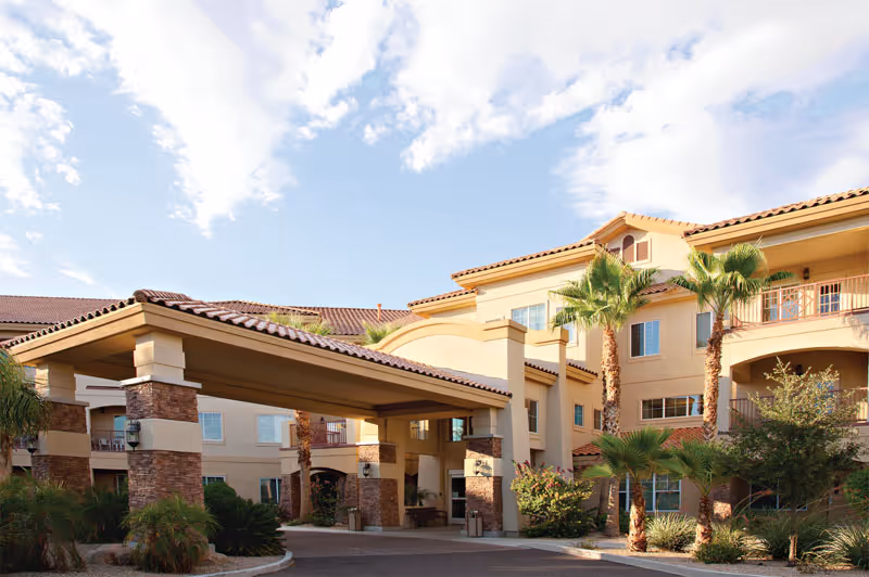 Exterior view of a multi-story senior living facility with a covered entrance, palm trees, and landscaping under a partly cloudy sky.