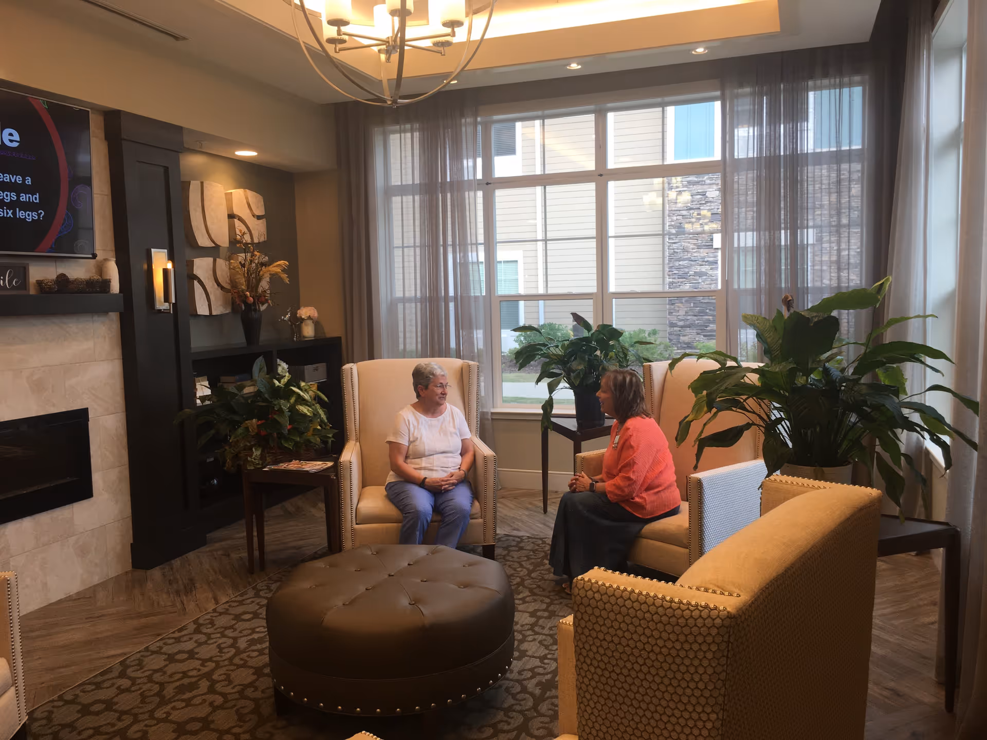 Two women sitting and talking in a well-lit living room area with large windows, beige armchairs, a round brown ottoman, indoor plants, and a fireplace with a TV mounted above it.
