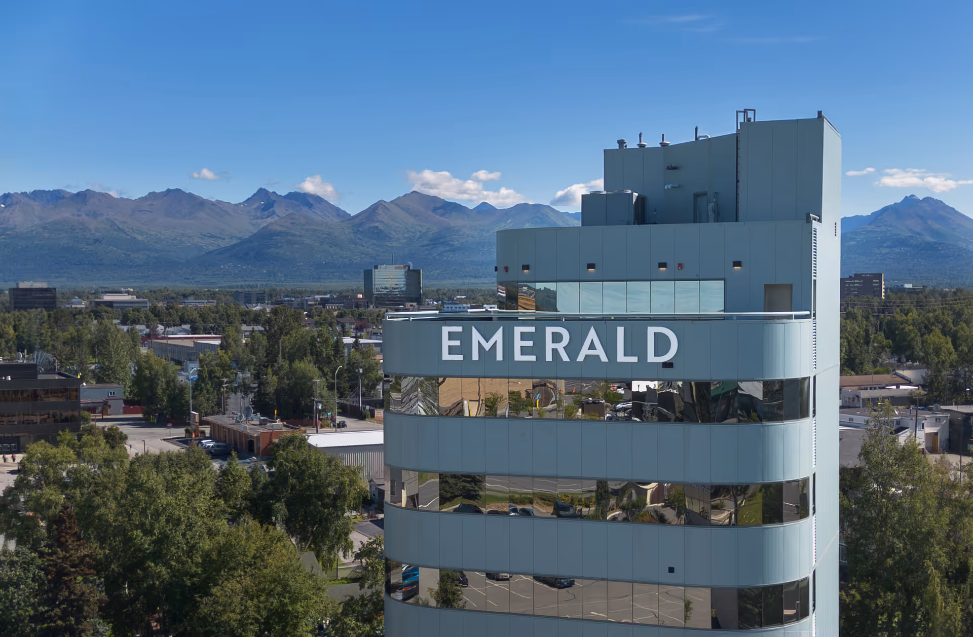 A tall modern building with reflective windows and the word 'EMERALD' displayed prominently near the top, surrounded by trees and other buildings with mountains in the background under a clear blue sky.
