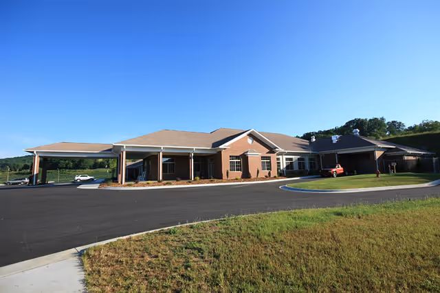 Exterior view of a single-story brick assisted living facility building with a covered entrance and a driveway. The building is surrounded by a grassy area and trees in the background under a clear blue sky.