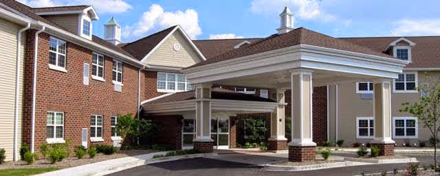 Exterior view of a senior living facility building with a covered entrance, brick and beige siding, multiple windows, and a clear blue sky with some clouds.