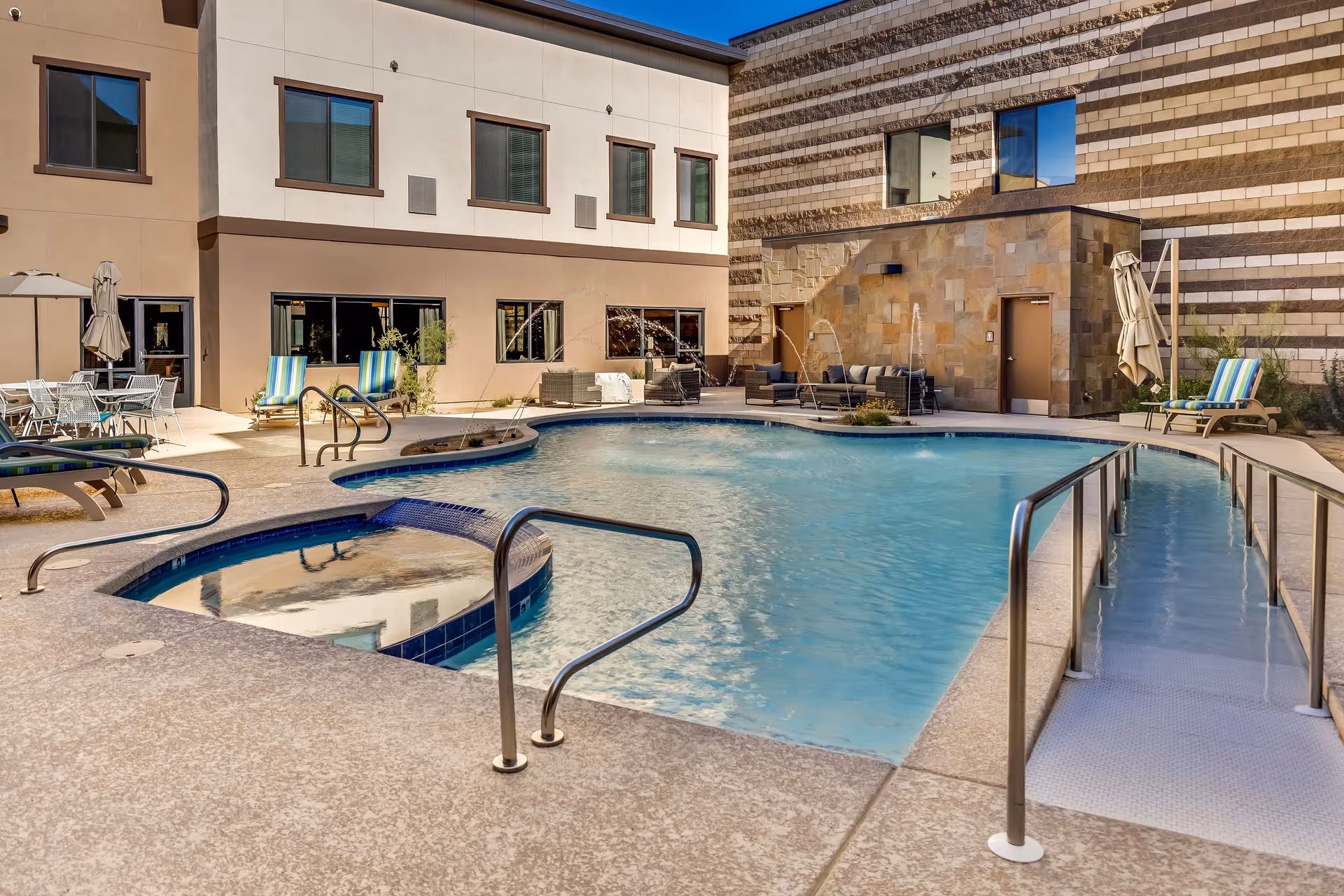 Outdoor swimming pool area at Legacy Village of Salt River with a hot tub, lounge chairs with striped cushions, patio tables with umbrellas, and water fountains. The pool has a ramp for accessibility and is surrounded by a beige textured deck. The building exterior features beige and brown tones with windows overlooking the pool.