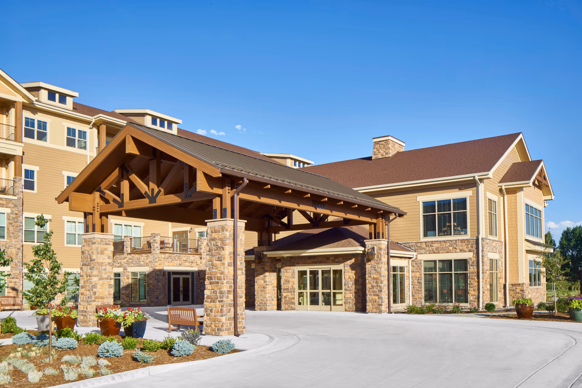 Exterior view of a senior living facility building with a large covered entrance supported by stone pillars. The building has beige siding with stone accents and multiple windows. There are landscaped plants and a clear blue sky in the background.