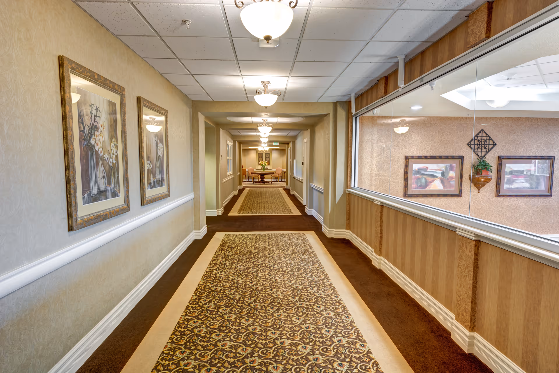 A long, carpeted interior hallway with a patterned runner, framed artwork, ceiling lights, and a large interior window overlooking a seating area.