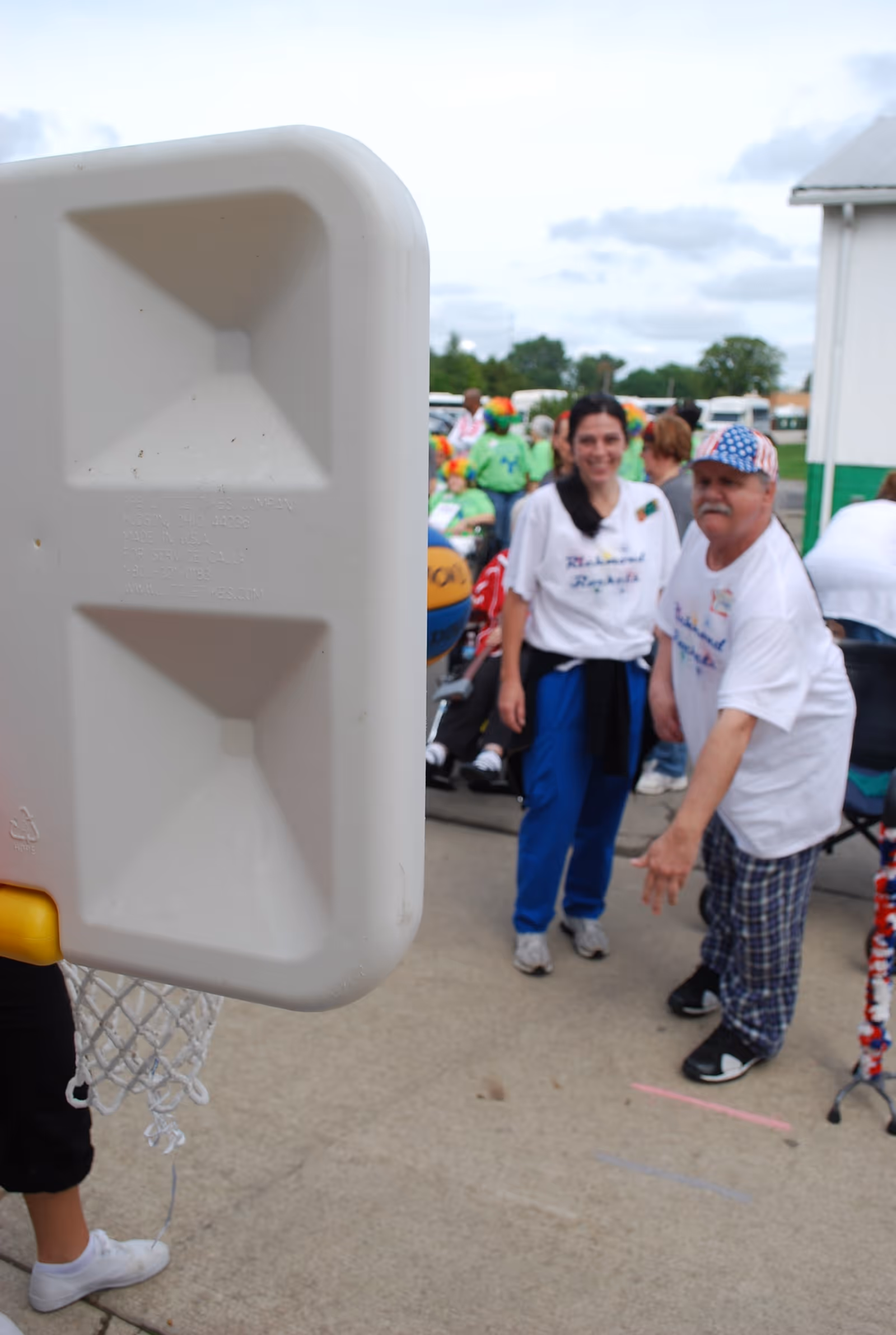An outdoor scene at Medilodge of Richmond showing a man and a woman participating in a basketball game. The man is in mid-throw aiming at a plastic basketball hoop, while the woman stands nearby smiling. Other people and greenery are visible in the background.