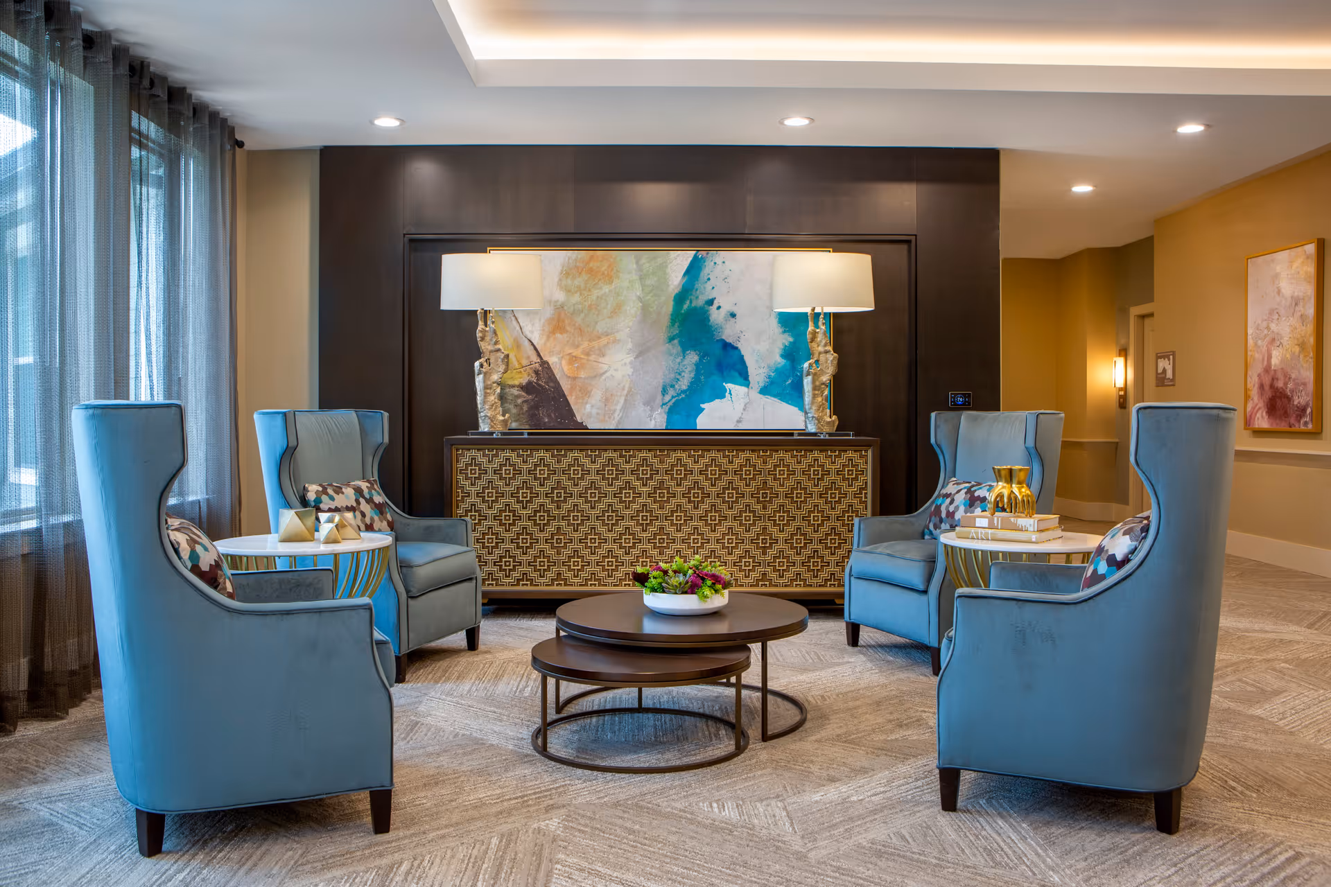 A cozy seating area with four blue upholstered armchairs arranged around two nested round coffee tables with a small plant centerpiece. Behind the chairs is a decorative sideboard with a geometric pattern, topped with two matching lamps and a large abstract painting. The room has large windows with sheer curtains on the left and warm-toned walls with artwork on the right.