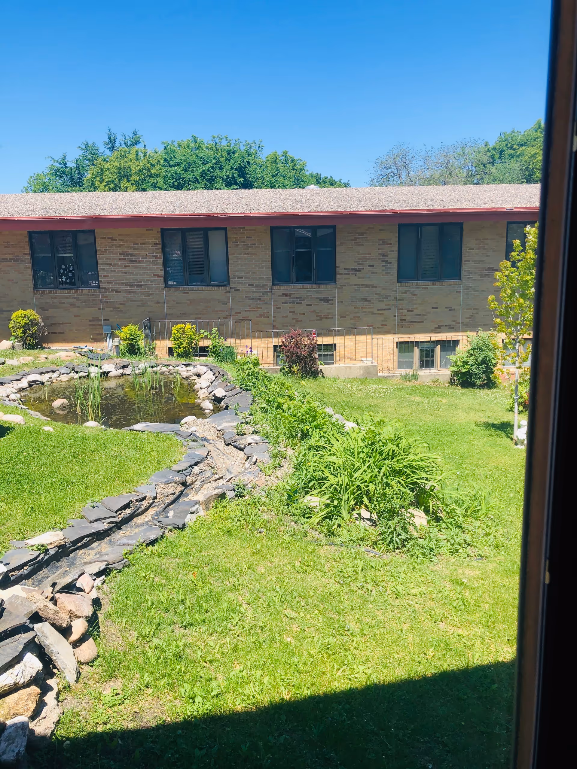 View through a window of a grassy courtyard with a small pond and stone-lined water feature in front of a brick senior living building.