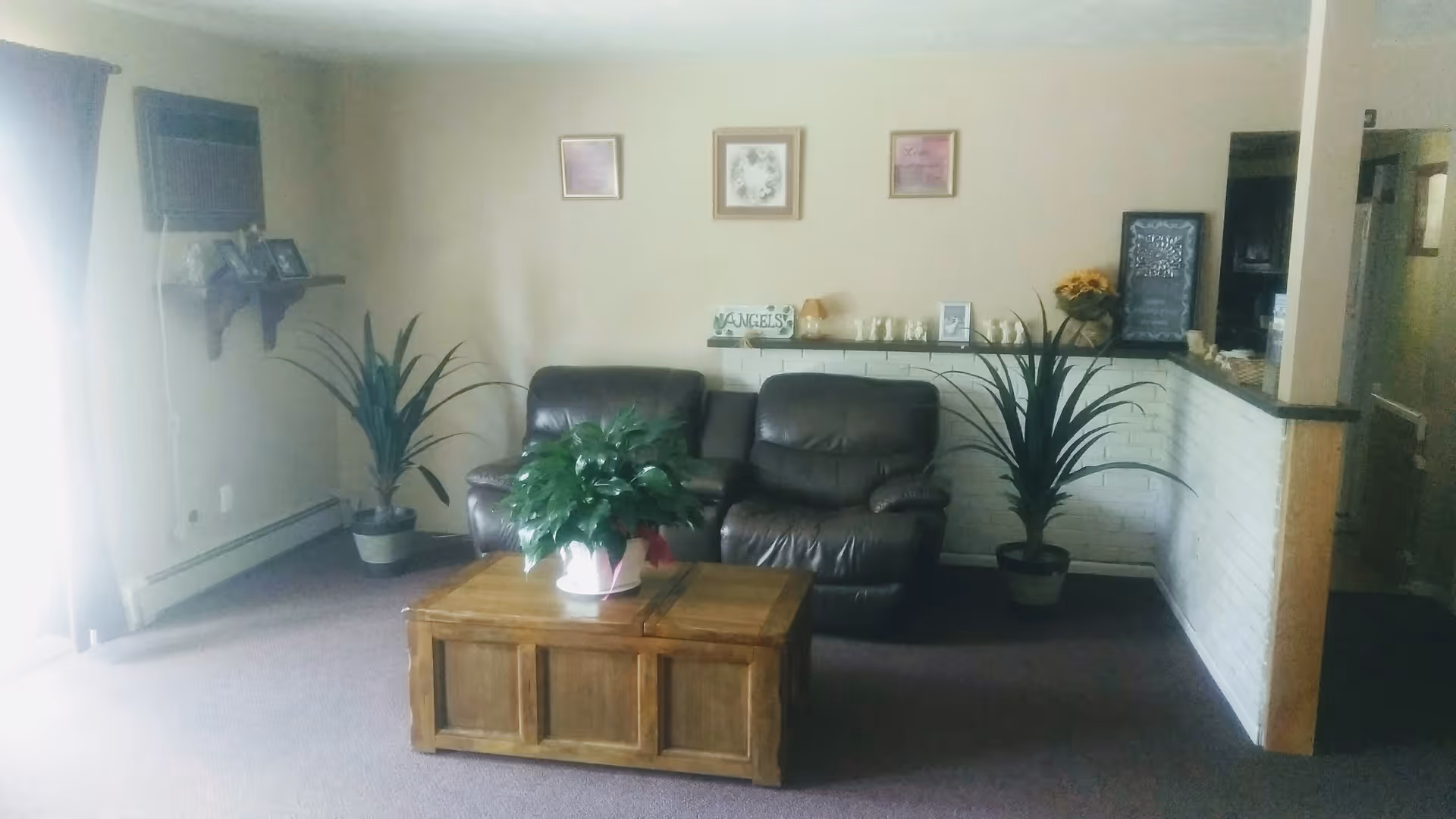 A cozy living room area with two dark leather recliners, a wooden coffee table with a potted plant on it, two large potted plants on either side of the recliners, and a white brick half-wall with decorative items and framed pictures on the wall above.