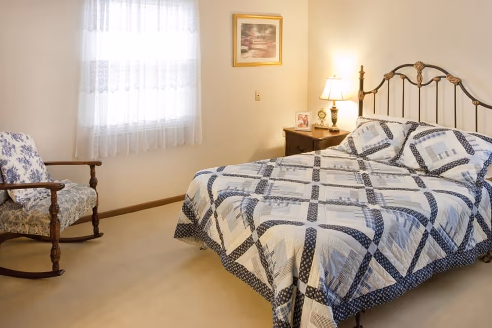 A cozy bedroom with a metal-framed bed covered by a blue-and-white quilt, a nightstand with a lamp, and a rocking chair by a lace-curtained window.