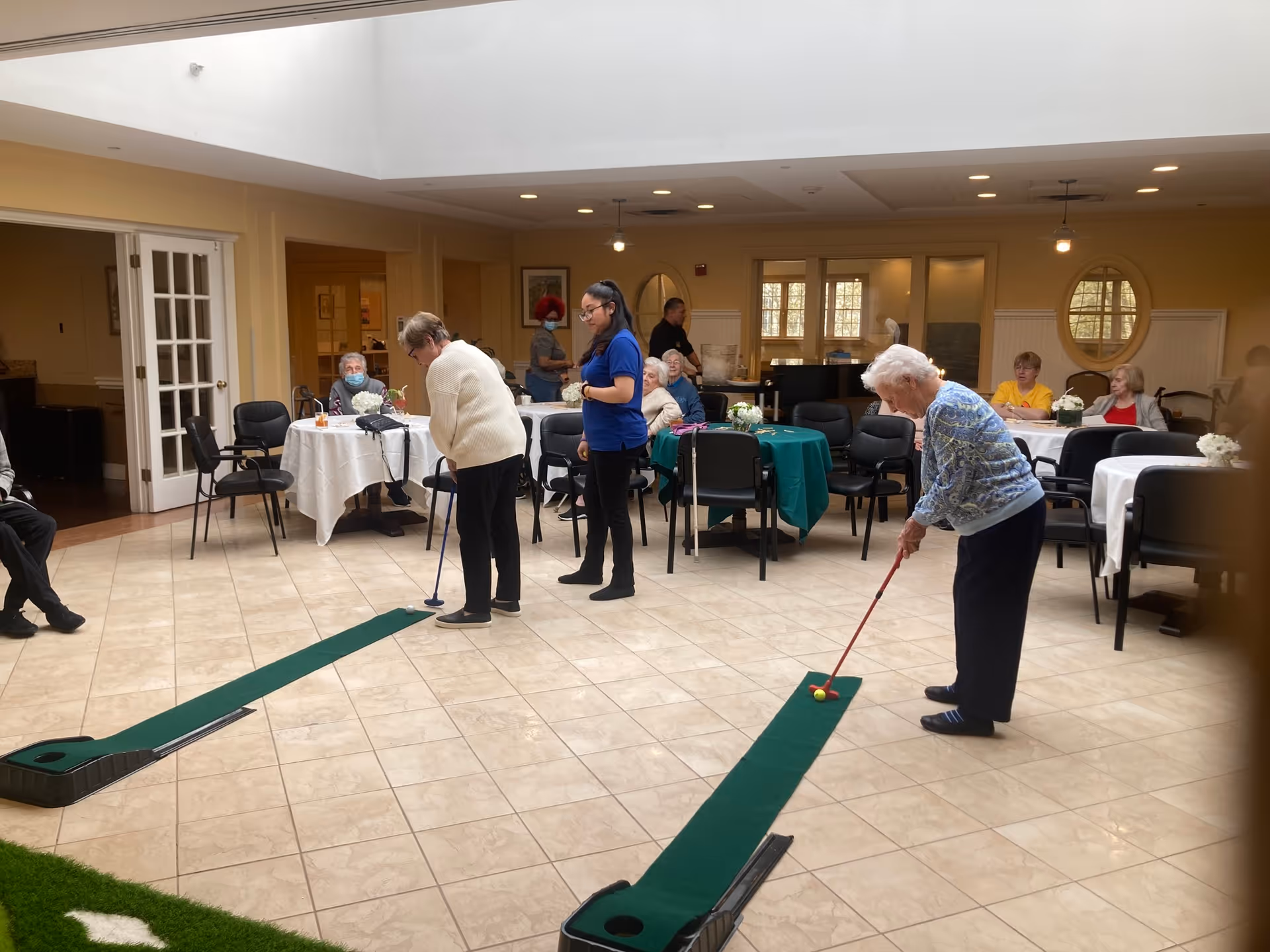 A group of elderly people and a caregiver in a spacious, well-lit common area with tiled floors and tables covered with white and green tablecloths. Two elderly women are playing indoor mini-golf on green putting mats while others sit at tables watching or chatting.