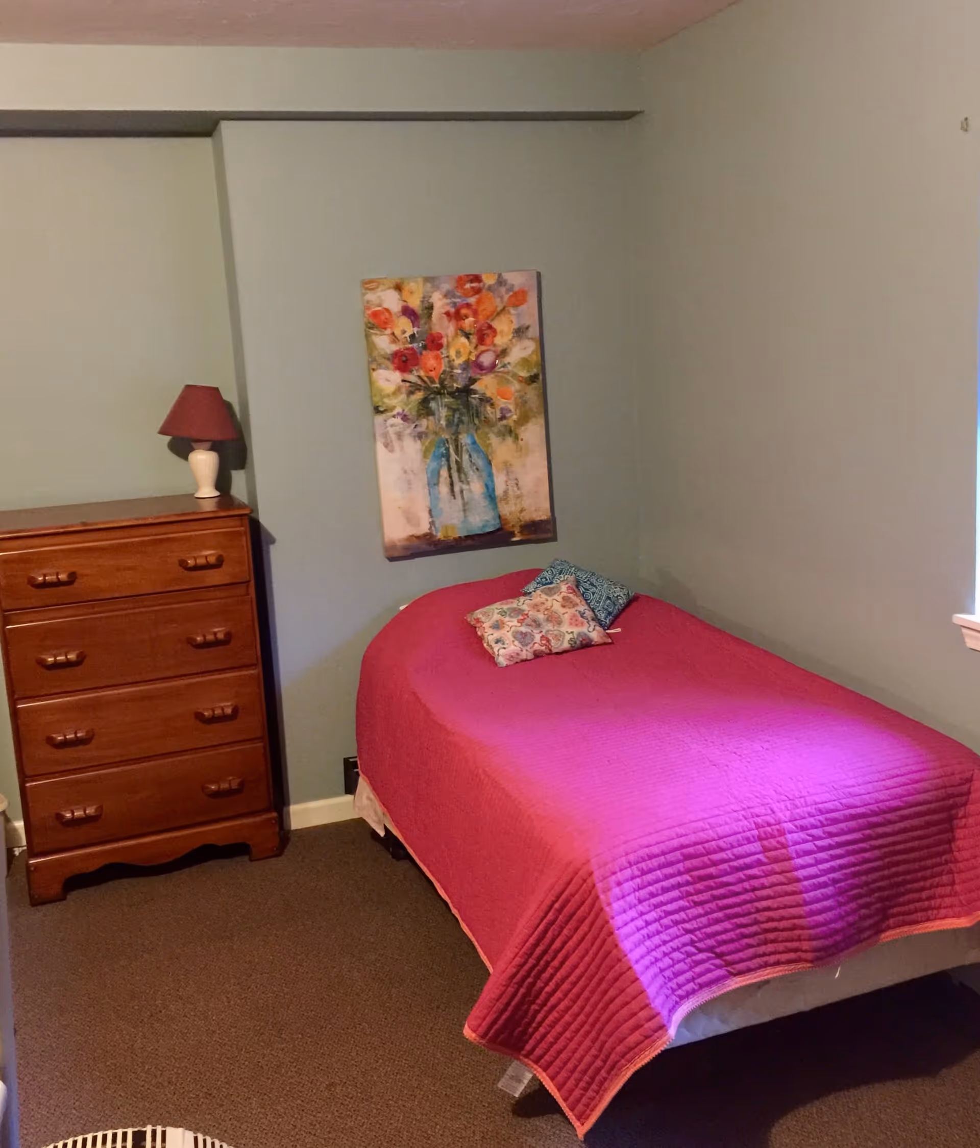 A small bedroom with a single bed covered in a bright pink quilt and two decorative pillows. Next to the bed is a wooden chest of drawers with a small lamp on top. A colorful painting of flowers in a vase hangs on the wall above the bed. The walls are painted light green and there is a window on the right side.