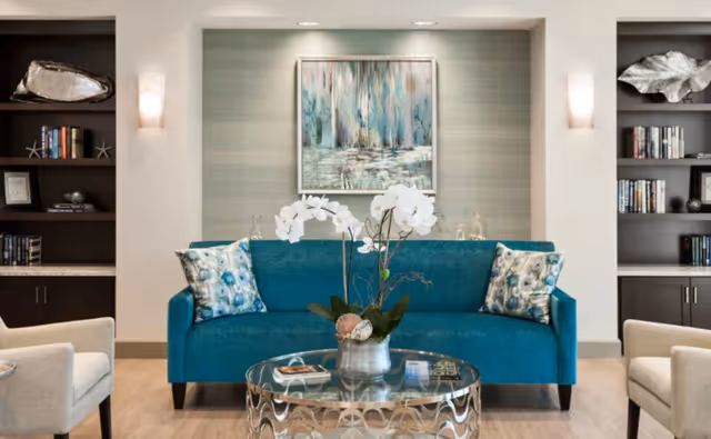 A teal sofa in a bright seating area with a glass coffee table topped with a white orchid, framed art on the wall, and built-in bookcases on either side.