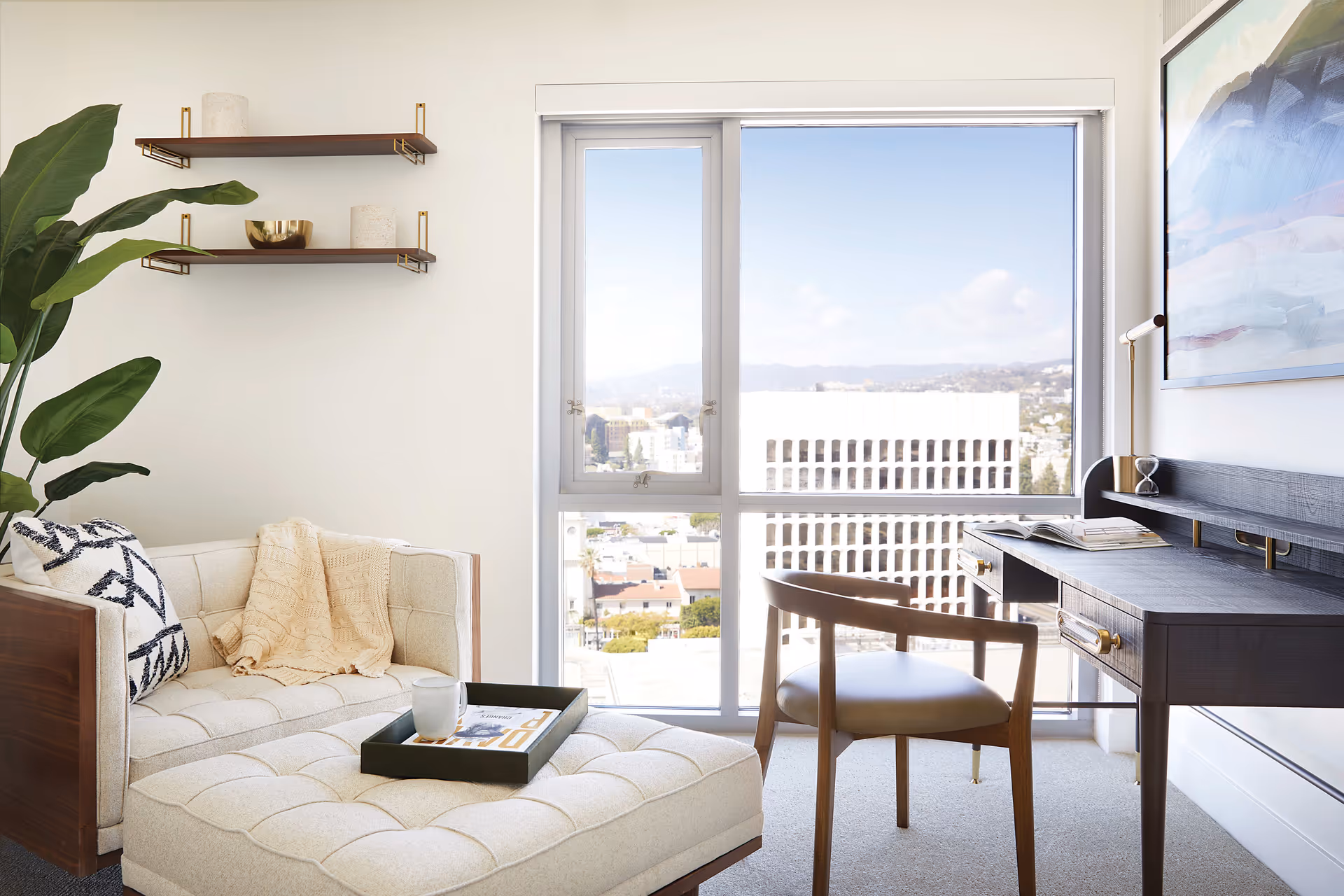 Bright living room with a tufted chaise lounge, wooden desk and chair beside a large window overlooking the city.