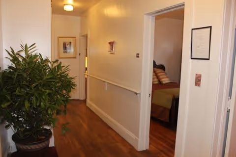 A hallway with wooden flooring and cream-colored walls featuring a handrail on one side. There is a large potted plant on the left side near the entrance of the hallway. On the right side, an open door reveals a bedroom with a bed that has a brown headboard and pillows with striped pillowcases. A framed picture hangs on the wall at the end of the hallway.