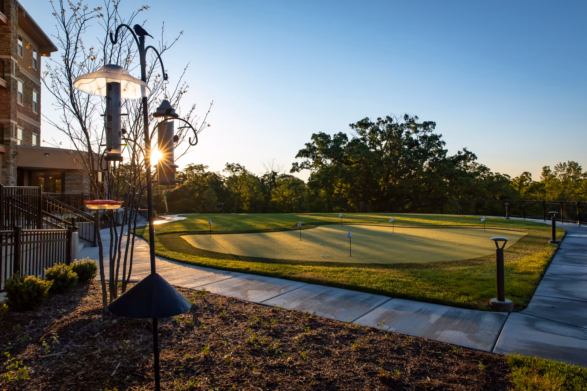 Outdoor putting green at a senior living facility with several small flags on the green, surrounded by a paved walkway. Bird feeders and a small tree are in the foreground, and a multi-story building is visible on the left. The sun is low in the sky, casting a warm light over the scene.