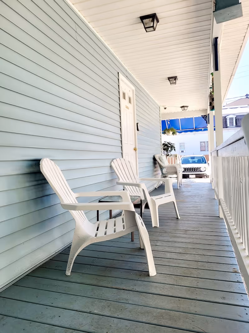 Covered front porch with white plastic chairs and a small table along a light-blue sided building overlooking a parked car.