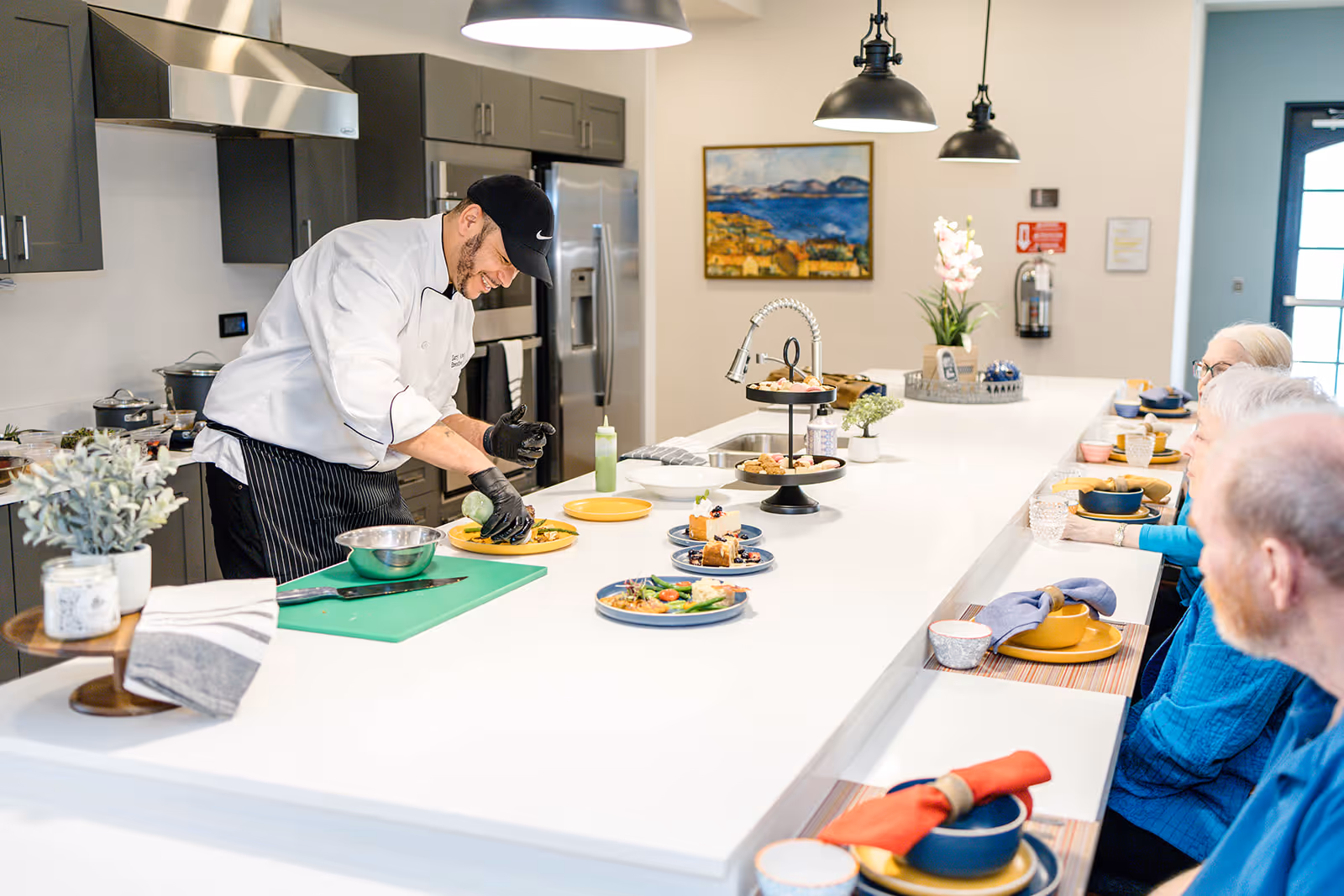A chef wearing a white coat and black cap is preparing and plating food on a long white kitchen island. Several elderly people are seated on the opposite side of the island, watching the chef. The kitchen has modern appliances, pendant lights, and a painting on the wall in the background.