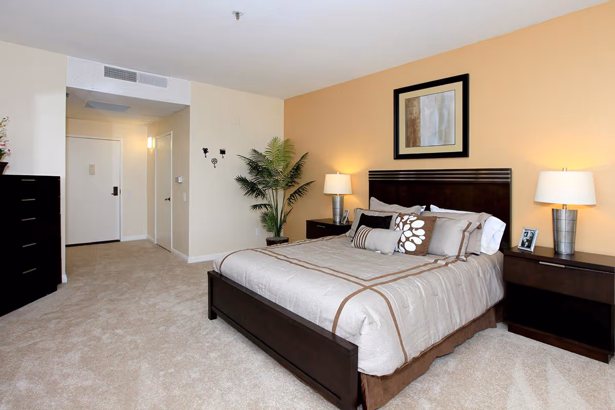 A neatly arranged bedroom with a large bed featuring beige and brown bedding and multiple pillows. On either side of the bed are dark wood nightstands with lamps and a small framed photo. A framed abstract painting hangs above the headboard on a light brown accent wall. The room has beige carpet and walls, a tall green plant in the corner, a dark wood dresser on the left, and a white door leading to another room or hallway.