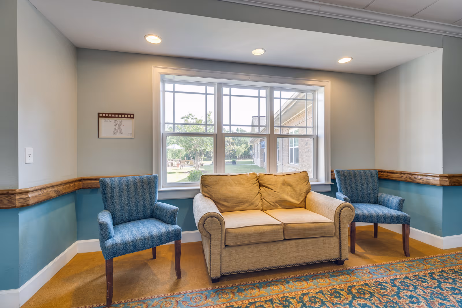 A cozy sitting area inside Chatham Ridge Assisted Living featuring a beige loveseat flanked by two blue upholstered armchairs. The seating is positioned in front of a large window that lets in natural light and offers a view of the outdoor garden and building exterior. The walls are painted light gray with a teal lower section and wooden chair rail. A patterned rug covers the floor beneath the furniture.