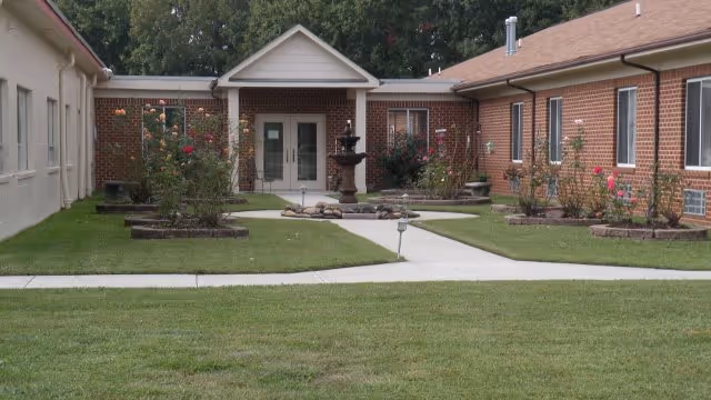 View of the exterior courtyard area of Pine View Assisted Living Facility featuring a central walkway leading to a double-door entrance. The courtyard has a grassy lawn, flower beds with blooming roses, and a multi-tiered water fountain in the center. The building is made of brick with several windows and a pitched roof.