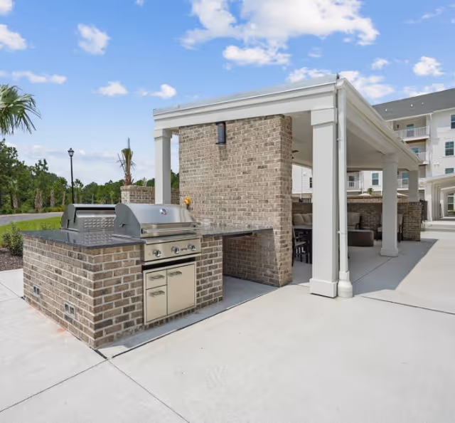 Outdoor grilling area with a stainless steel barbecue grill built into a brick counter. The area is adjacent to a covered patio with seating, supported by white columns. The background shows a clear blue sky with some clouds and a multi-story residential building.