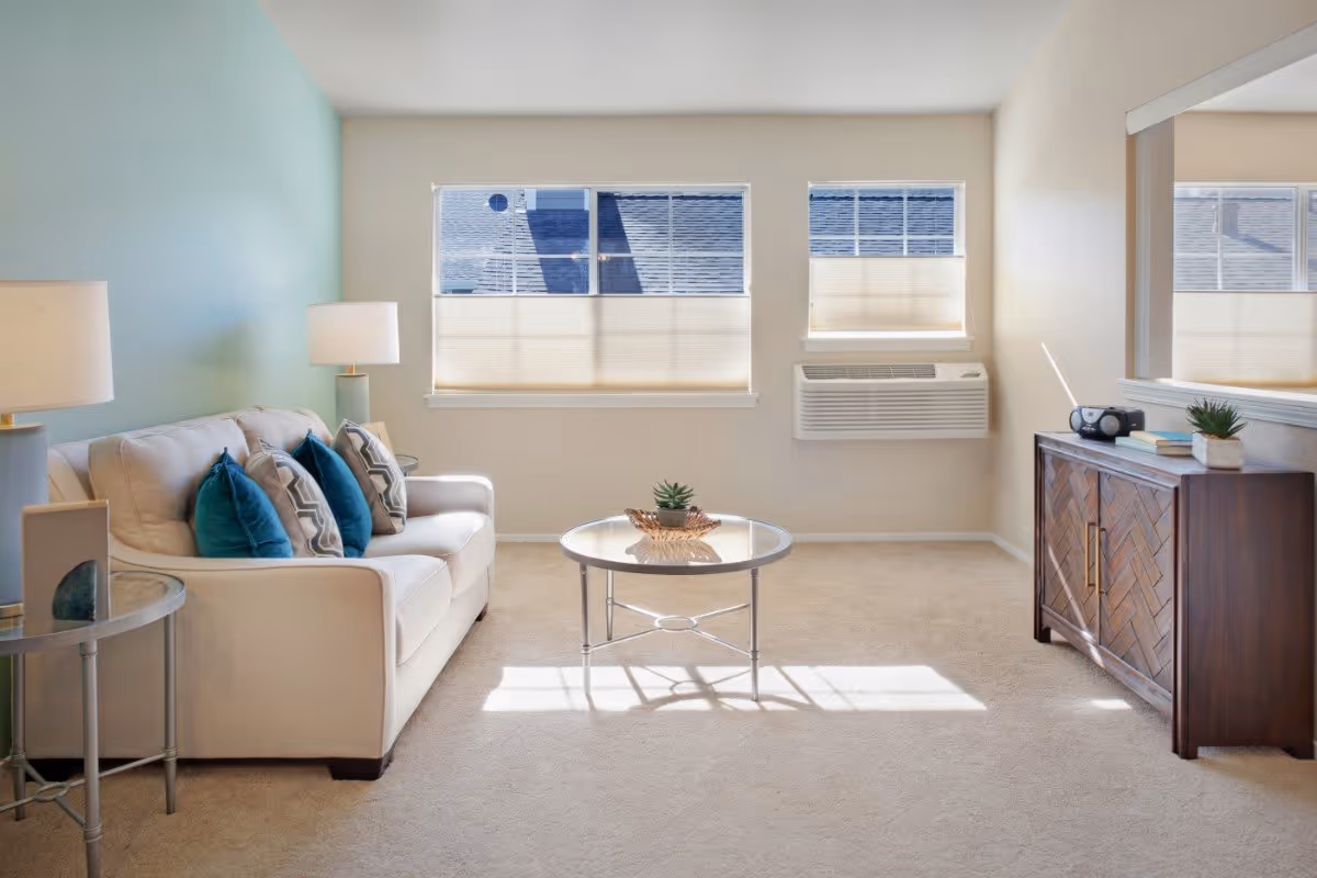A bright living room with a beige sofa adorned with blue and patterned throw pillows, two side tables with lamps, a round glass coffee table with a small plant centerpiece, a wooden cabinet with a radio and books, and three windows with blinds partially closed.