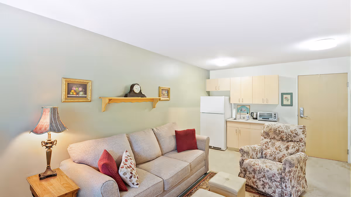 A cozy living area in a senior living facility featuring a beige sofa with red and patterned cushions, a floral armchair, a wooden side table with a decorative lamp, and a small kitchen area with a refrigerator, cabinets, a toaster oven, and a kettle. The walls are light green and white, decorated with framed pictures and a clock on a wooden shelf.