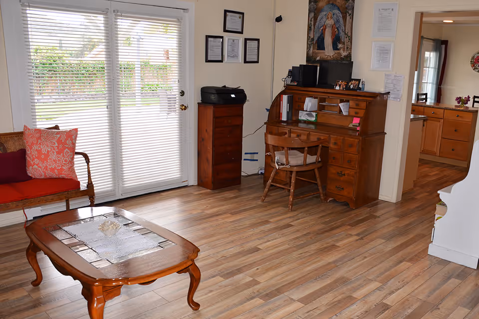 A cozy living room area with wooden flooring, a wooden coffee table with a lace runner, a wooden bench with red and patterned cushions, a wooden roll-top desk with a chair, and a tall wooden cabinet with a printer on top. French doors with blinds lead outside, and there are framed certificates and a religious painting on the wall.