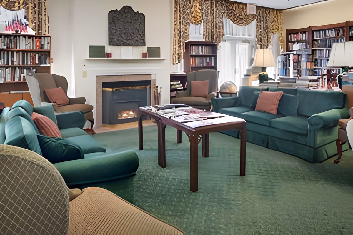 A cozy living room with green sofas and armchairs arranged around a wooden coffee table. The room features a lit fireplace, bookshelves filled with books, large windows with patterned curtains, and table lamps providing warm lighting.