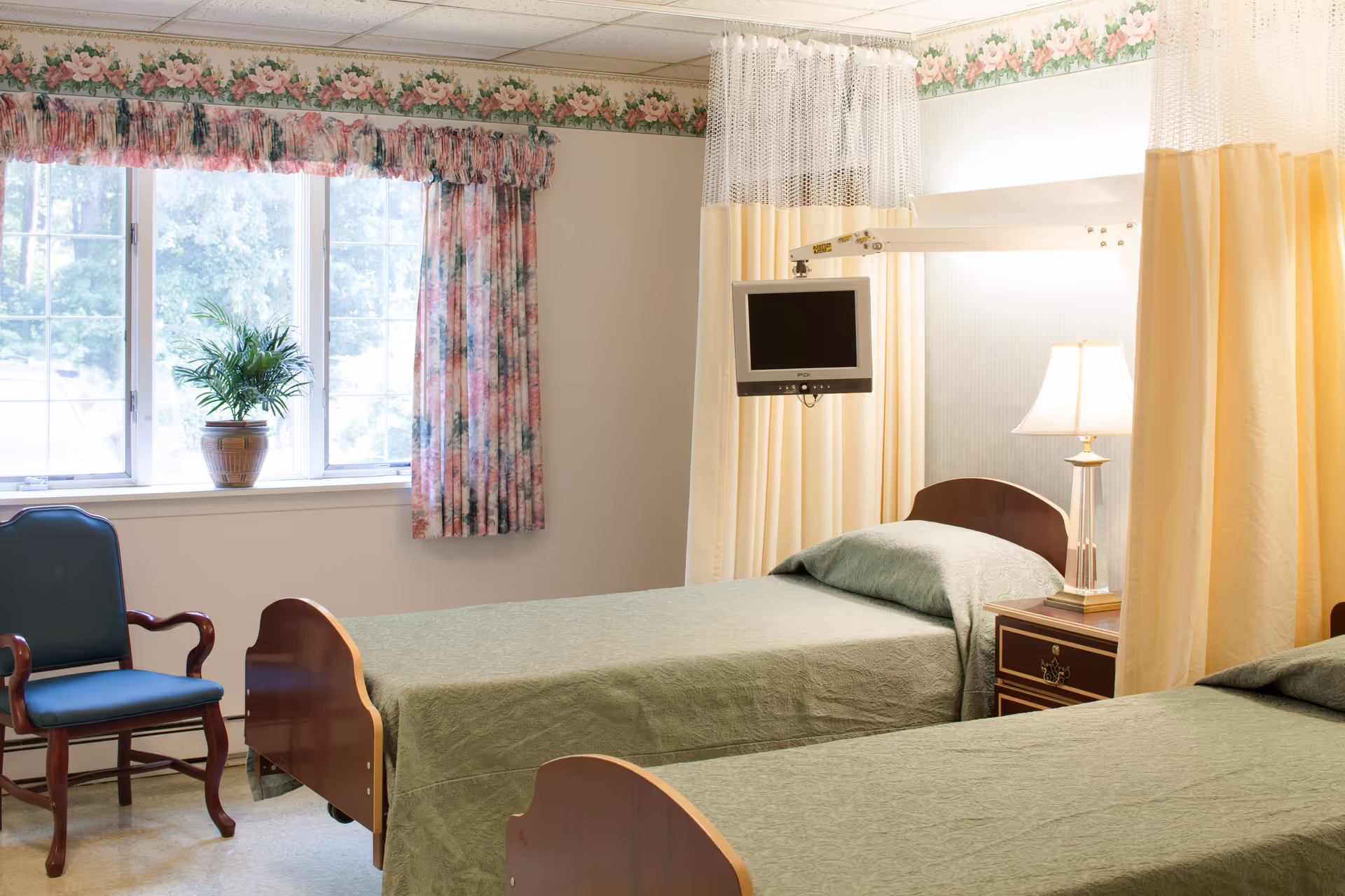 A tidy shared nursing room with two beds, a bedside table and lamp, a wall-mounted TV, and a window with floral curtains.