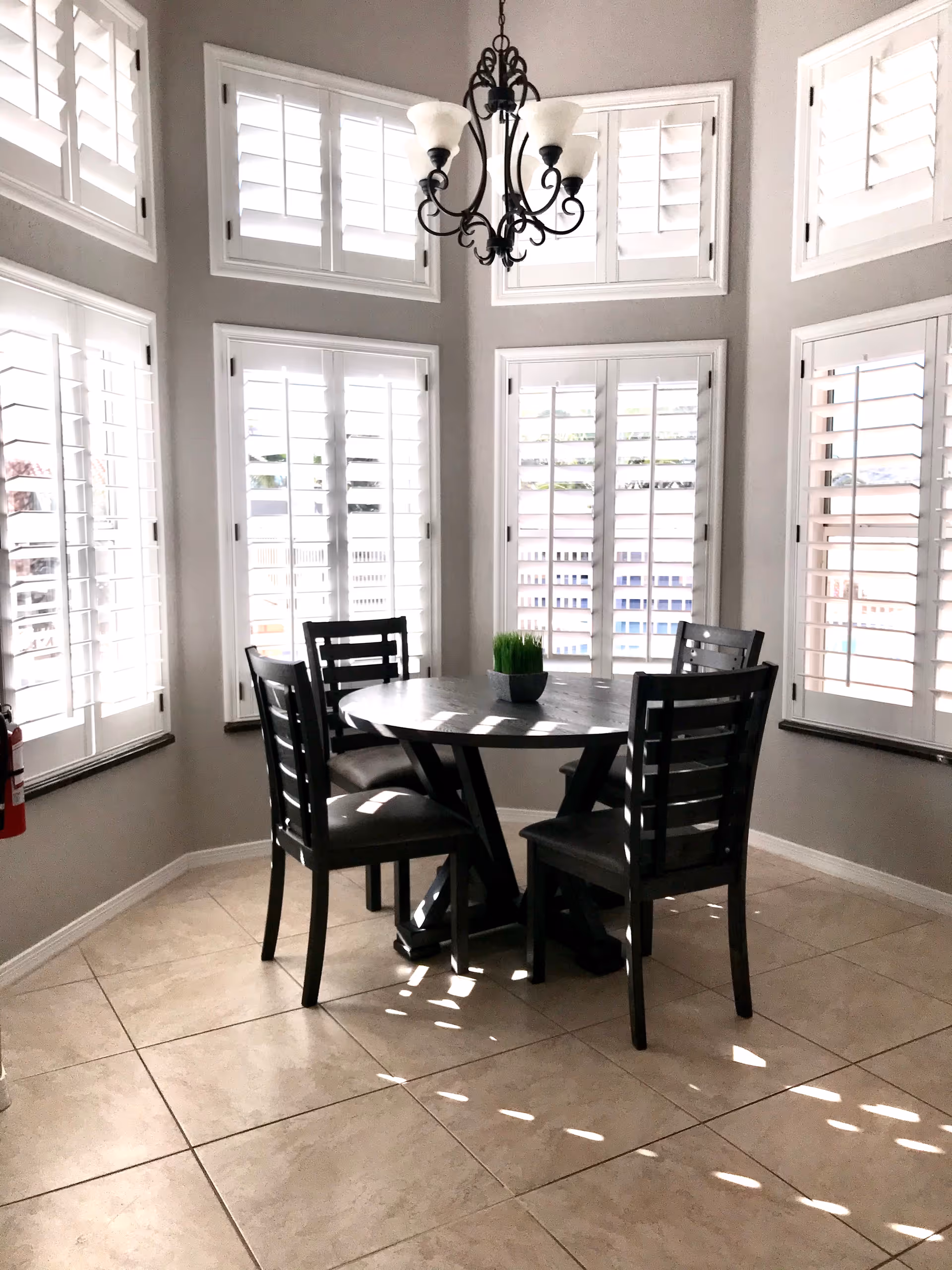 A bright dining area with a round wooden table surrounded by four dark wooden chairs. The room has large windows with white plantation shutters allowing sunlight to stream in, casting shadows on the tiled floor. A black chandelier with white lampshades hangs above the table, and a small green plant is placed in the center of the table.