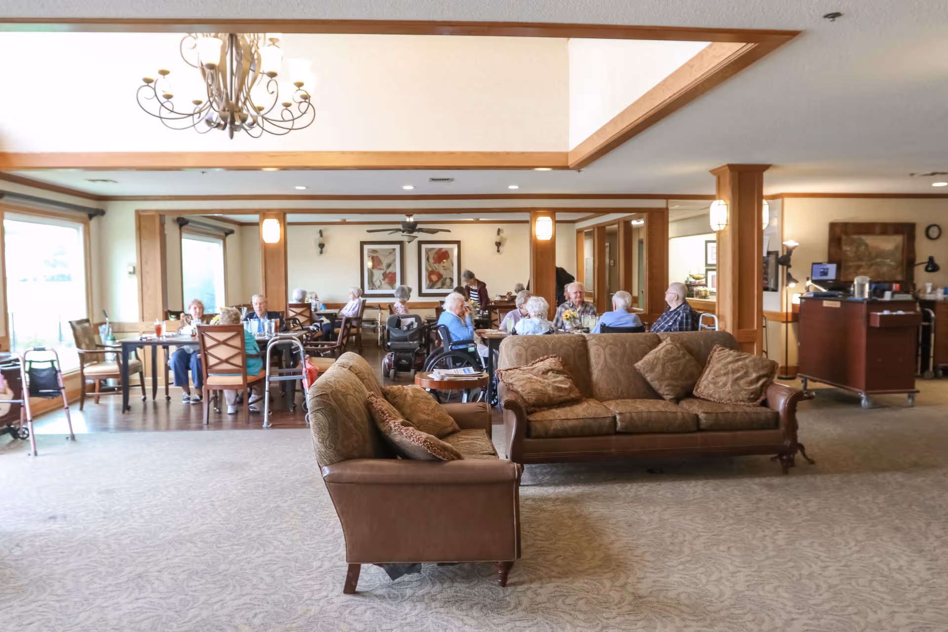 A spacious common area in a senior living facility with two brown sofas in the foreground and several elderly residents sitting at tables in the background. The room has large windows, wooden trim, a chandelier, and framed artwork on the walls.