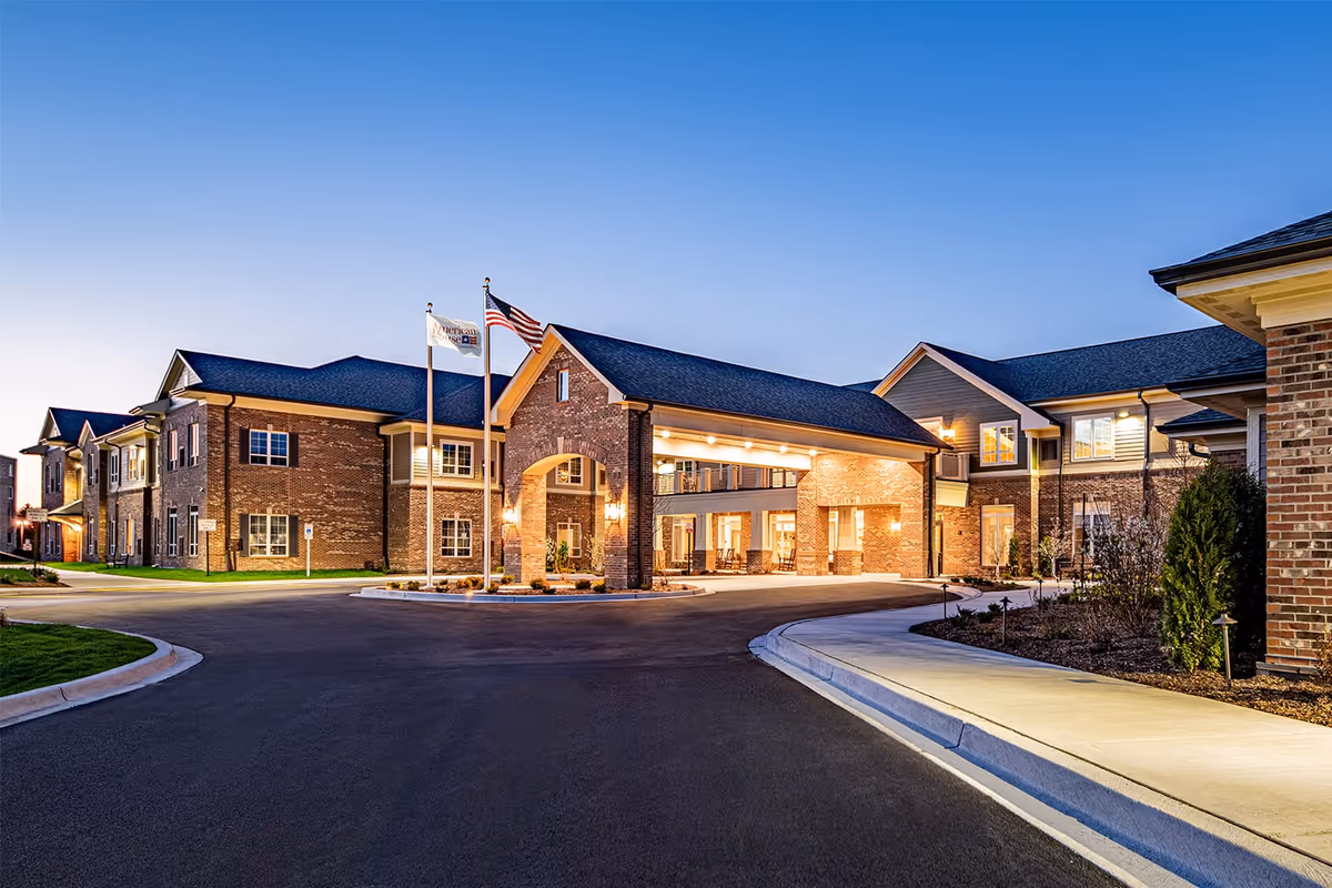 Exterior view of the American House Mount Prospect senior living facility at dusk, showing a large brick building with a covered entrance, two flagpoles with flags, well-lit windows, and a paved driveway with landscaped areas.