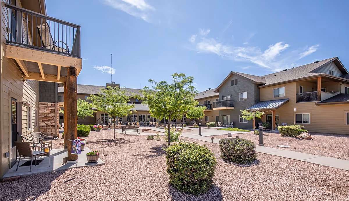 Outdoor courtyard area of Majestic Rim Retirement Living featuring a gravel ground, small trees, bushes, benches, and a paved walkway surrounded by multi-story residential buildings under a clear blue sky.