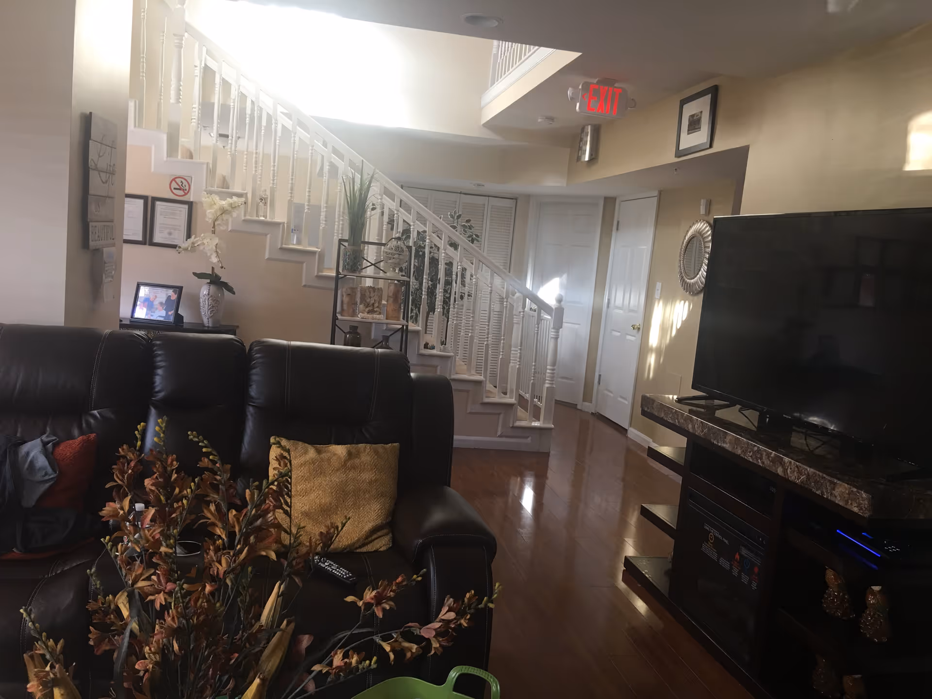 Interior view of a living room in an assisted living facility with a dark leather couch adorned with pillows, a TV on a stand with a marble top, a staircase with white railings leading to an upper floor, and decorative plants and framed pictures on the walls.