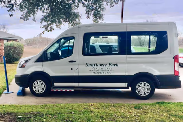 A white passenger van parked on a driveway with the text Sunflower Park Health Care and a phone number printed on the side. The van is under a tree with green grass in the foreground and a building partially visible on the left.