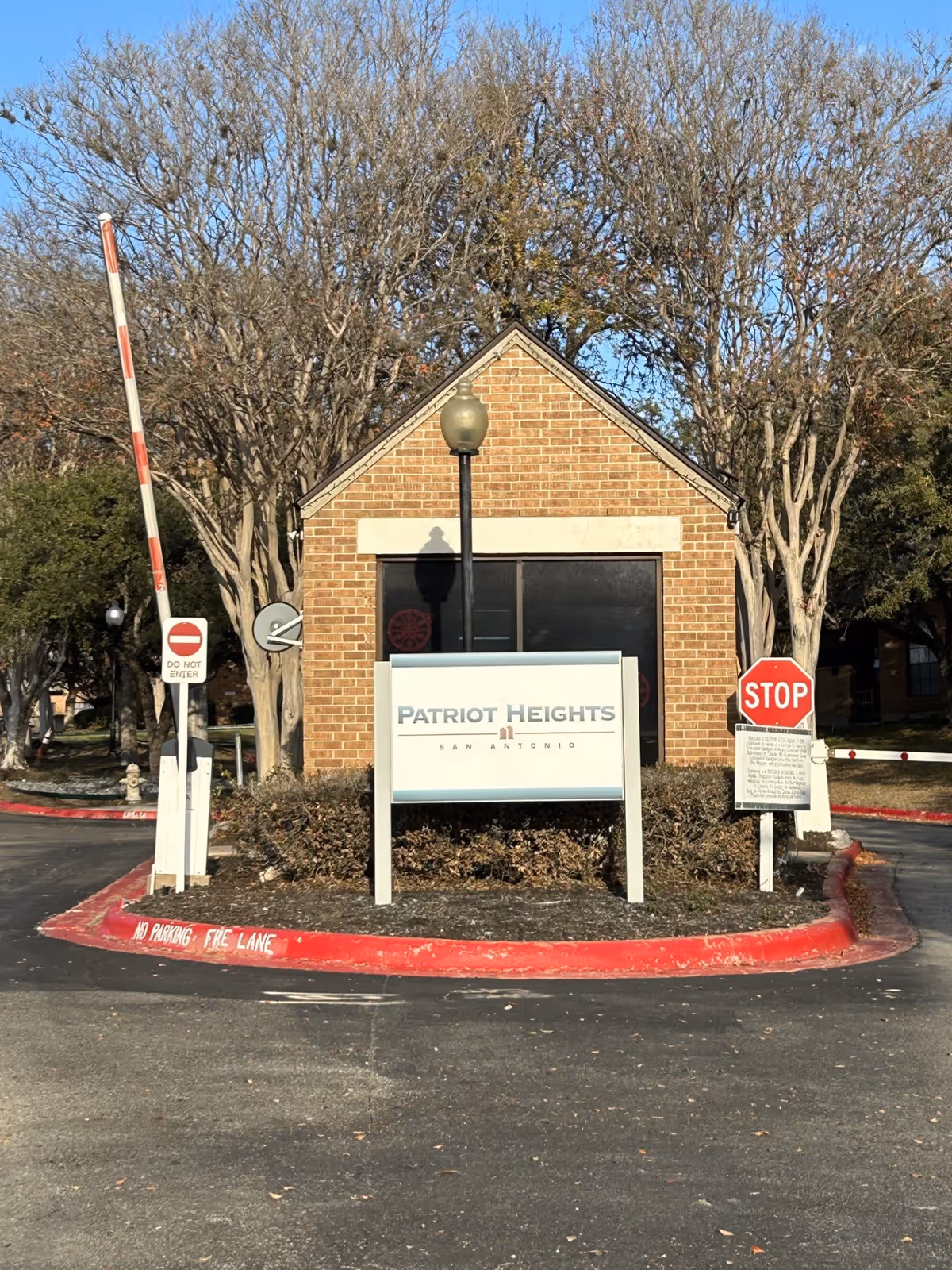 Entrance gatehouse of Patriot Heights facility with a brick building, a sign reading 'Patriot Heights San Antonio', a stop sign, a do not enter sign, and a red and white striped barrier arm. Trees without leaves are visible in the background.